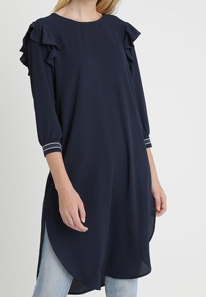 Frau trägt eine marineblaue Langarm-Tunika mit gerafften Schultern, gestreiften Manschettendetails und Seitenschlitzen über hellblauen Jeans.