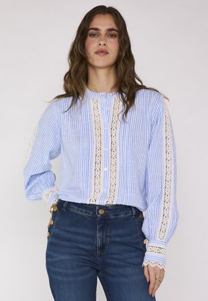 Donna che indossa una blusa a righe blu e bianche con dettagli in pizzo, abbinata a jeans blu scuro a vita alta con bottoni laterali.