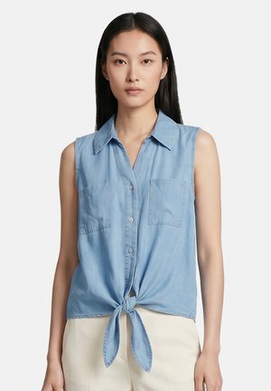 Giovane donna con lunghi capelli neri che indossa una camicia azzurra senza maniche, abbottonata e annodata in vita, e pantaloni chiari.