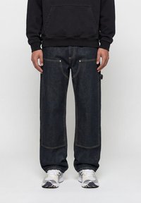 Άτομο που φοράει σκούρο μπλε φαρδιά τζιν παντελόνια με εμφανείς ραφές, συνδυασμένα με λευκά και γκρι αθλητικά παπούτσια και μαύρο πουλόβερ.