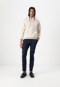 Beige hættetrøje med frontlomme, parret med marineblå slim-fit bukser og sorte loafers. Simpelt design, blød tekstur og neutrale farver.