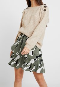 Vrouw in een beige gebreide trui met knoopdetails op de schouders, gecombineerd met een groen, wit en zwart camouflagepatroon flared rok.