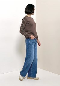 Chemise marron à manches longues avec des détails de couture latérale, associée à un jean bleu à jambes larges et des baskets de couleur claire. Fond uni.