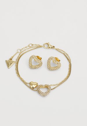 Conjunto de joyería en color dorado que incluye pendientes en forma de corazón y una pulsera, adornados con brillantes de strass, eslabones de cadena y un charm con logo.