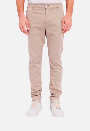 Uomo che indossa pantaloni slim beige chiaro e sneaker bianche con lacci, in piedi davanti a uno sfondo semplice.