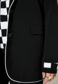 Černá blazer s bílým lemováním a zapínáním na jeden knoflík; má malou kapsu a je doplněna černobílým kostkovaným manžetem.