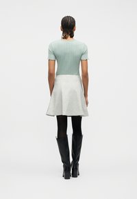 Γυναίκα με πλεξουδάκια στα μαλλιά, φοράει ανοιχτό πράσινο ριμπ τοπ με κοντά μανίκια, ανοιχτό γκρι φούστα με καμπάνα, μαύρες καλτσόν και μαύρες μπότες μέχρι το γόνατο.