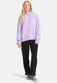 Sudadera con capucha lavanda, con bolsillo canguro, cordones ajustables y puños acanalados, combinada con pantalones de pana negros y zapatillas deportivas.