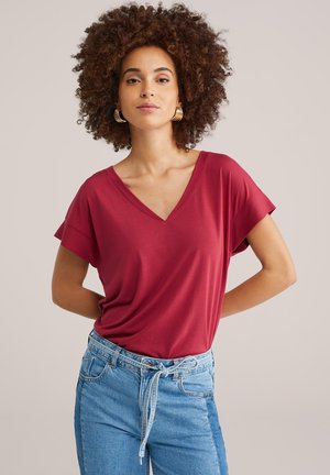 Moteris su garbanotais plaukais, dėvinti auksinius žiedinius auskarus, raudoną V formos trumpomis rankovėmis megztinį ir aukštu liemeniu mėlynas džinsus su surišta diržu.