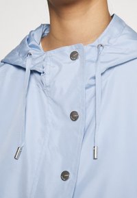 Vaaleansininen sadetakki, jossa on huppu, hopeiset napit ja säätönyörit. Suuri, kiiltävä kangas rentoa istuvuutta.