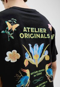 Černé bavlněné tričko s barevnou grafikou květin a ptáků. Text zní "ATELIER ORIGINALS" a "ZALOŽENO V ROCE 1990."