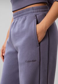 Χέρι στην τσέπη μωβ φόρμας Calvin Klein με ελαστική μέση και μαύρες κάθετες ρίγες στο πλάι.