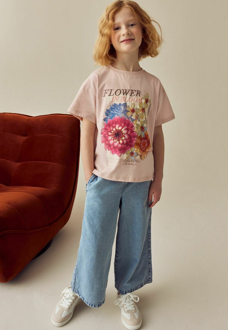 Child with curly red hair stands smiling wearing a pink flower print shirt, wide jeans, and white sneakers beside a brown chair.