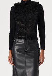 Kvinde iført en sort ribstrikket trøje med lynlås og imiteret pels foran samt en sort læder pencilskørt, hænderne afslappet ned langs siden.