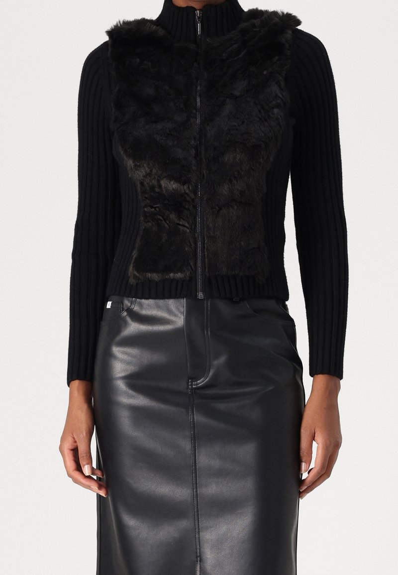 Kvinde iført en sort ribstrikket trøje med lynlås og imiteret pels foran samt en sort læder pencilskørt, hænderne afslappet ned langs siden.