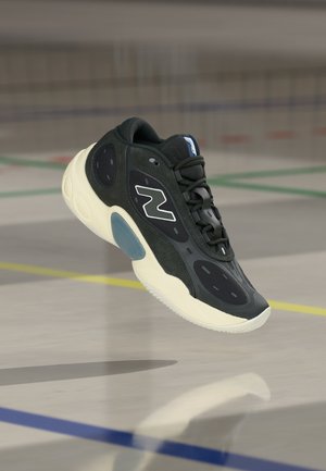 Czarno-biały sportowy trampki z amortyzowaną podeszwą i logo "N", ukazany w powietrzu nad podłogą krytego boiska sportowego.