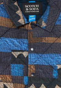 Quiltet jakke med blå, brun og svart abstrakt geometrisk mønster, svarte trykknapper og "Scotch & Soda Amsterdam"-etikett inni kragen.