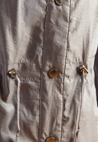 Giacca beige chiaro realizzata in tessuto testurizzato, con bottoni dorati, lacci e una vestibilità rilassata con dettagli di cucitura visibili.
