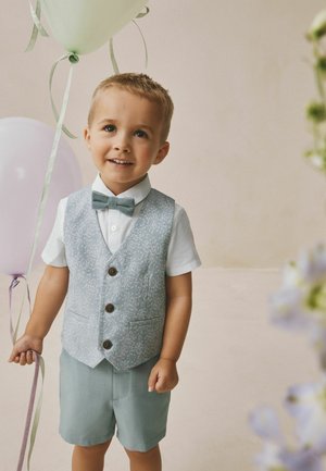 Jonge jongen die lacht, gekleed in een lichtblauw vest met patroon, vlinderdas en korte broek, houdt pastelkleurige ballonnen vast tegen een neutrale achtergrond.
