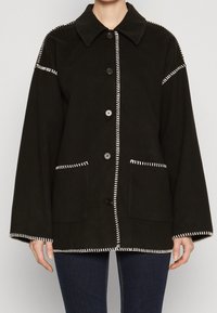 Czarna kurtka z wywiniętym kołnierzem, z białym przeszyciem, dwoma przednimi kieszeniami i zapięciem na guziki. Wykonana z miękkiego materiału.