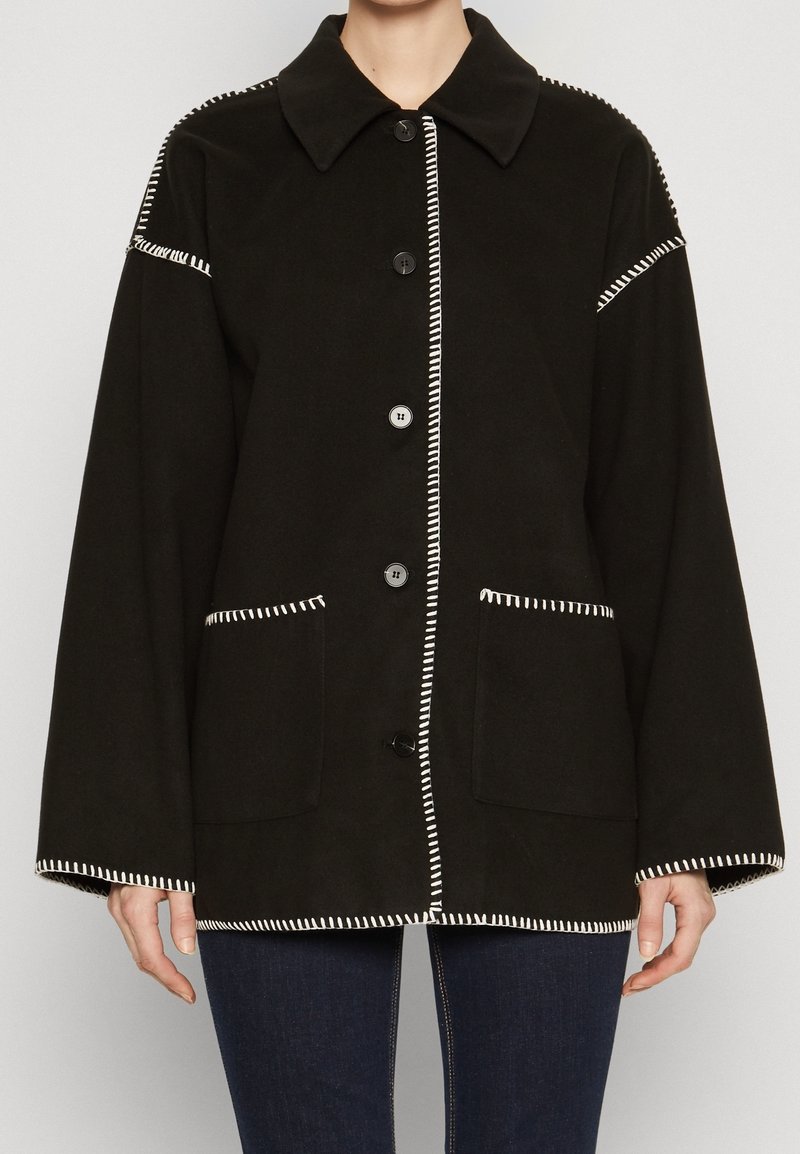 Czarna kurtka z wywiniętym kołnierzem, z białym przeszyciem, dwoma przednimi kieszeniami i zapięciem na guziki. Wykonana z miękkiego materiału.