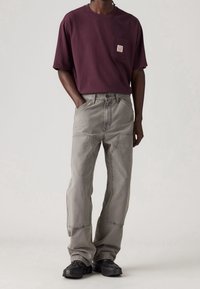 Camiseta de algodón burdeos con bolsillo en el pecho y etiqueta, combinada con pantalones vaqueros holgados de color gris claro. El conjunto se completa con zapatillas negras.