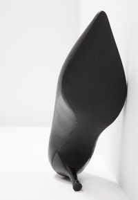 Chaussure à talon haut en cuir noir reposant sur son talon contre un mur blanc, montrant la semelle pointue et le talon aiguille fin.