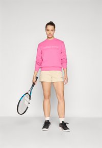 Rosa sweatshirt med hvit logo, beige shorts, svarte sportsko, og holder et tennisrack. Glatt stoff og atletisk passform.