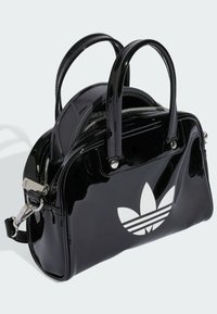 Sac à main en cuir verni noir, de forme arrondie, avec deux poignées, quincaillerie en métal argenté, fermeture zippée et détail du logo trefoil blanc d'Adidas.