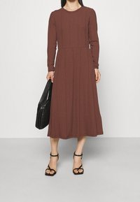 Długo rękawowa, prążkowana sukienka midi w kolorze brązowym, z okrągłym dekoltem i dopasowanym górnym krojem, który rozszerza się od talii. Połączona z czarną torbą.