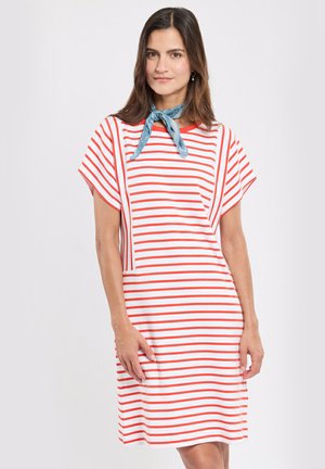 Femme portant une robe blanche à rayures rouges horizontales et un foulard bleu autour du cou, debout devant un fond clair uni.