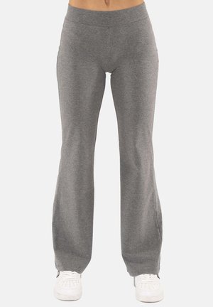 Donna con pantaloni tuta grigi svasati e sneakers bianchi, in piedi davanti a uno sfondo chiaro e uniforme.