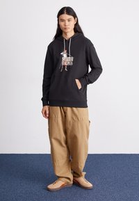 Black hooded sweatshirt with graphic print on front, kangaroo pocket, paired with light khaki baggy pants and tan sneakers.