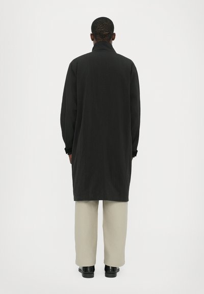 Long black coat with a high collar, made from a textured fabric. Features wide sleeves and is paired with light beige trousers and black shoes.
