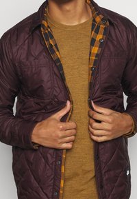 Burgundská prošívaná bunda přes hořčicový svetr, s kostkovanou podšívkou v žluté a černé barvě. Obsahuje přední kapsy a zapínání na knoflíky.