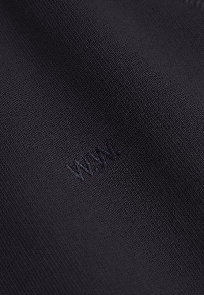 Približnji posnetek črne tkanine z nežno vezenim logotipom "W.N.W." v ujemajoči se črni niti.