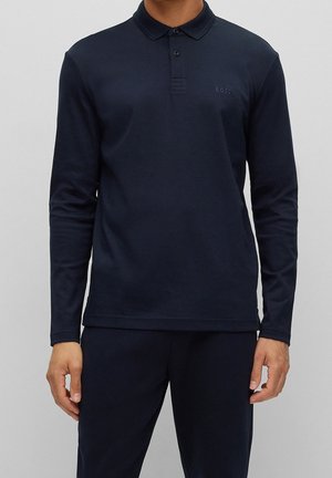 Man draagt een marineblauw poloshirt met lange mouwen en knopen, gecombineerd met een bijpassende marineblauwe broek, staand tegen een effen grijze achtergrond.