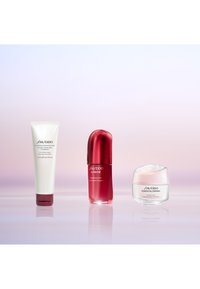 Drei Shiseido Hautpflegeprodukte auf einer reflektierenden Oberfläche mit einem sanften rosa-lila Farbverlauf im Hintergrund.