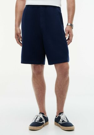 Mann trägt marineblaue Shorts und passende Sneakers mit weißen Schnürsenkeln und Sohlen, steht vor einem einfarbigen Hintergrund.