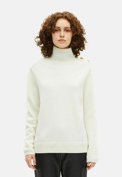 Femme aux cheveux bouclés portant un pull col roulé blanc avec des boutons dorés sur l'épaule et un pantalon noir, se tenant devant un fond uni.