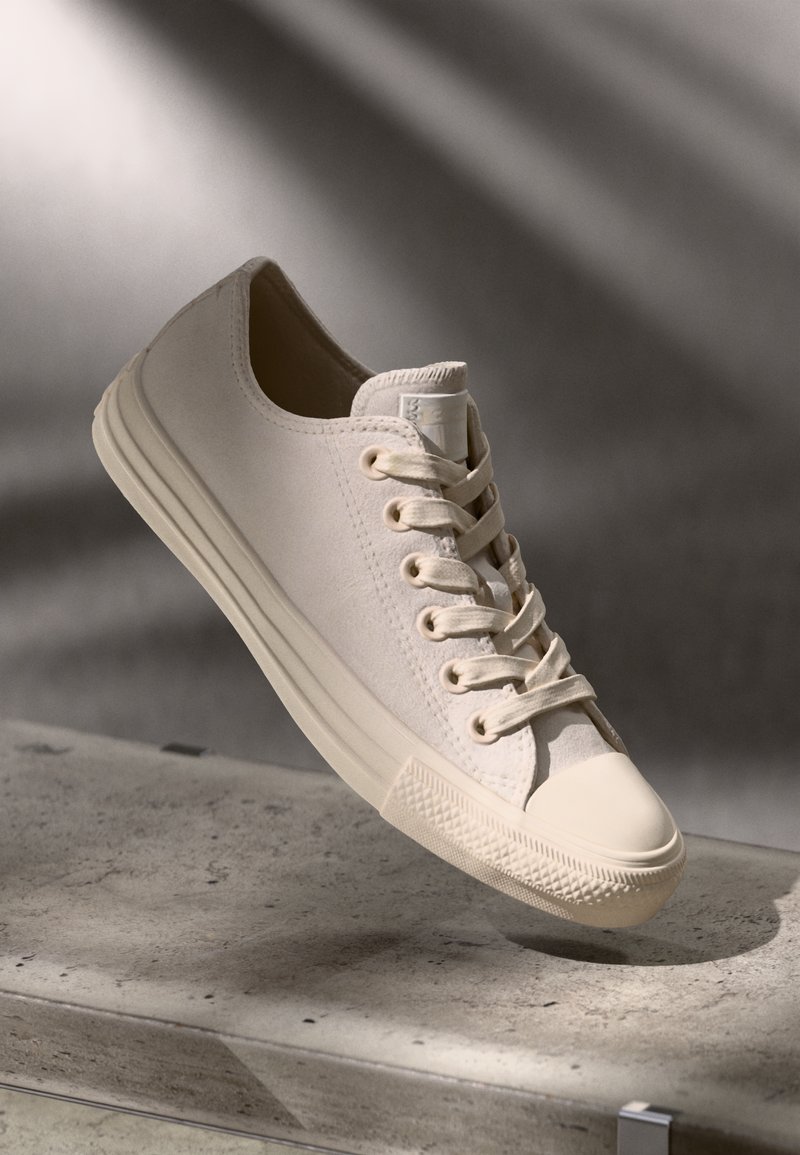 Zapatillas de lona beige con punta redondeada, suela de goma y seis ojales. Presentan una puntera texturizada y un parche de logo cosido en la lengüeta.