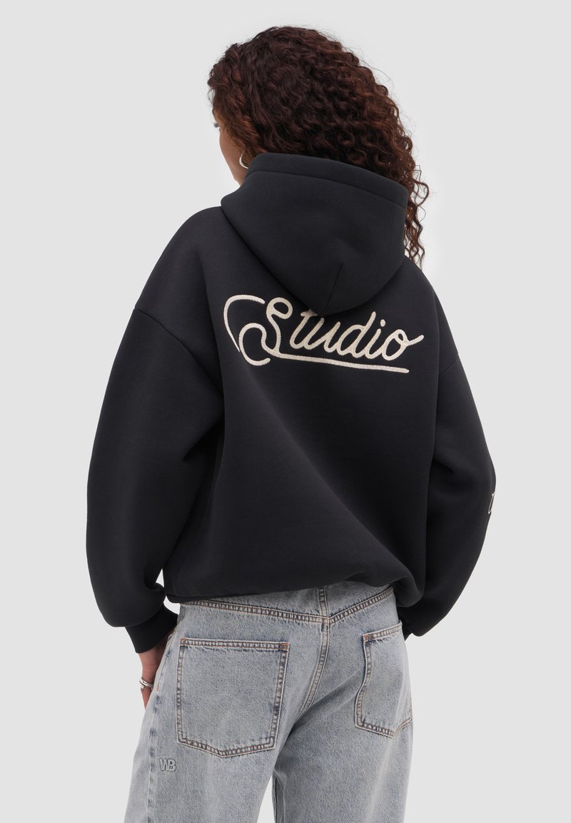 Kvinde med krøllet hår iført sort hættetrøje med teksten "Studio" på ryggen og lyseblå højttaljede jeans, der står mod en grå baggrund.