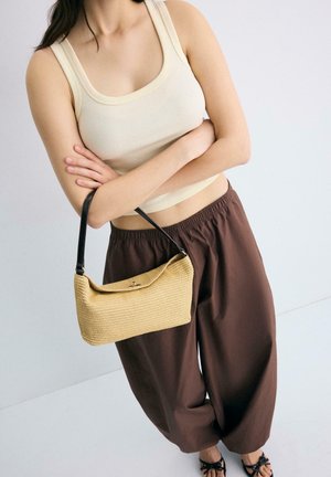 Mlada žena odjevena u kremasti top bez rukava i široke smeđe hlače, nosi malu bež pletenu torbu s crnim remenom, prekriženih ruku.