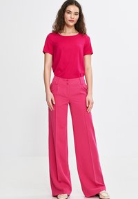 Fuksjowa bluzka z krótkim rękawem w połączeniu z fuksjowymi spodniami o wysokim stanie i szerokich nogawkach, z zapięciem na dwa guziki oraz fałdami z przodu. Gładka tekstura.
