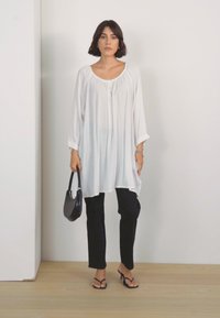 Witte lange mouwen tuniek met een ronde hals en gathered details, gecombineerd met een zwarte, strakke broek en zwarte flip-flops. Houdt een zwarte handtas vast.