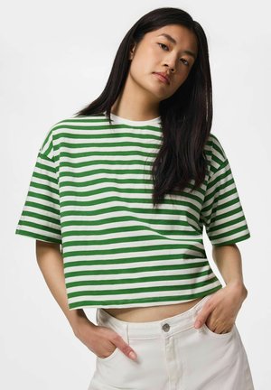 Jeune femme aux longs cheveux foncés portant un crop top à manches courtes rayé vert et blanc et un pantalon blanc, les mains dans les poches.