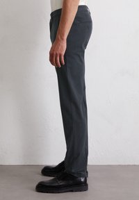 Herren Chinos in dunkelgrau, mit geradem Bein, aus einer Baumwollmischung. Mit schrägen Vorder- und einer knöpfbaren Gesäßtasche. Getragen mit schwarzen Schuhen.