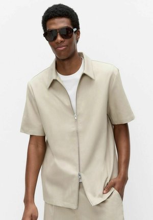 Man draagt een beige kortärmelig shirt met ritssluiting over een wit T-shirt, beige broek en grote donkere zonnebril, staand met één hand in zijn zak.