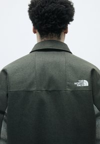 Zöld herringbone kabát, gallérral és fehér logóval a bal vállán. Texturált anyaggal és formázott sziluettel rendelkezik.