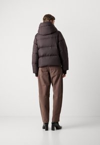 Personne portant une veste matelassée marron foncé avec capuche, un pantalon droit marron et des bottines noires à talons, se tenant contre un mur blanc uni.
